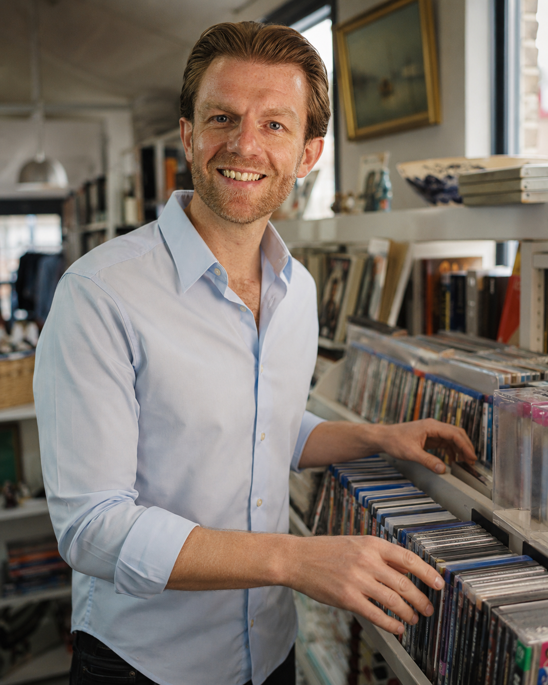 A smiling man with light brown hair and a beard, wearing a light blue button-down shirt, standing in a shop or library next to shelves filled with books and media cases.