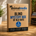 A brown cardboard box labeled 'animalmedia BLIND MYSTERY BOX OF BLU-RAYS' sits on a wooden table in a softly lit indoor setting with plants and books blurred in the background.