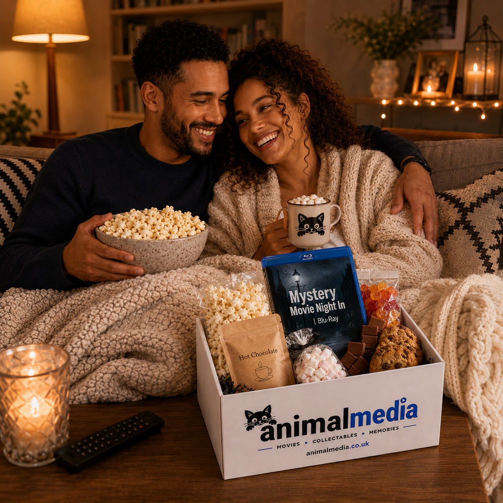 A man and woman sitting together on a couch under a cozy blanket, holding popcorn and a mug in a warmly lit living room. In front of them is an Animalmedia gift box filled with snacks, chocolates, marshmallows, and a Blu-ray.