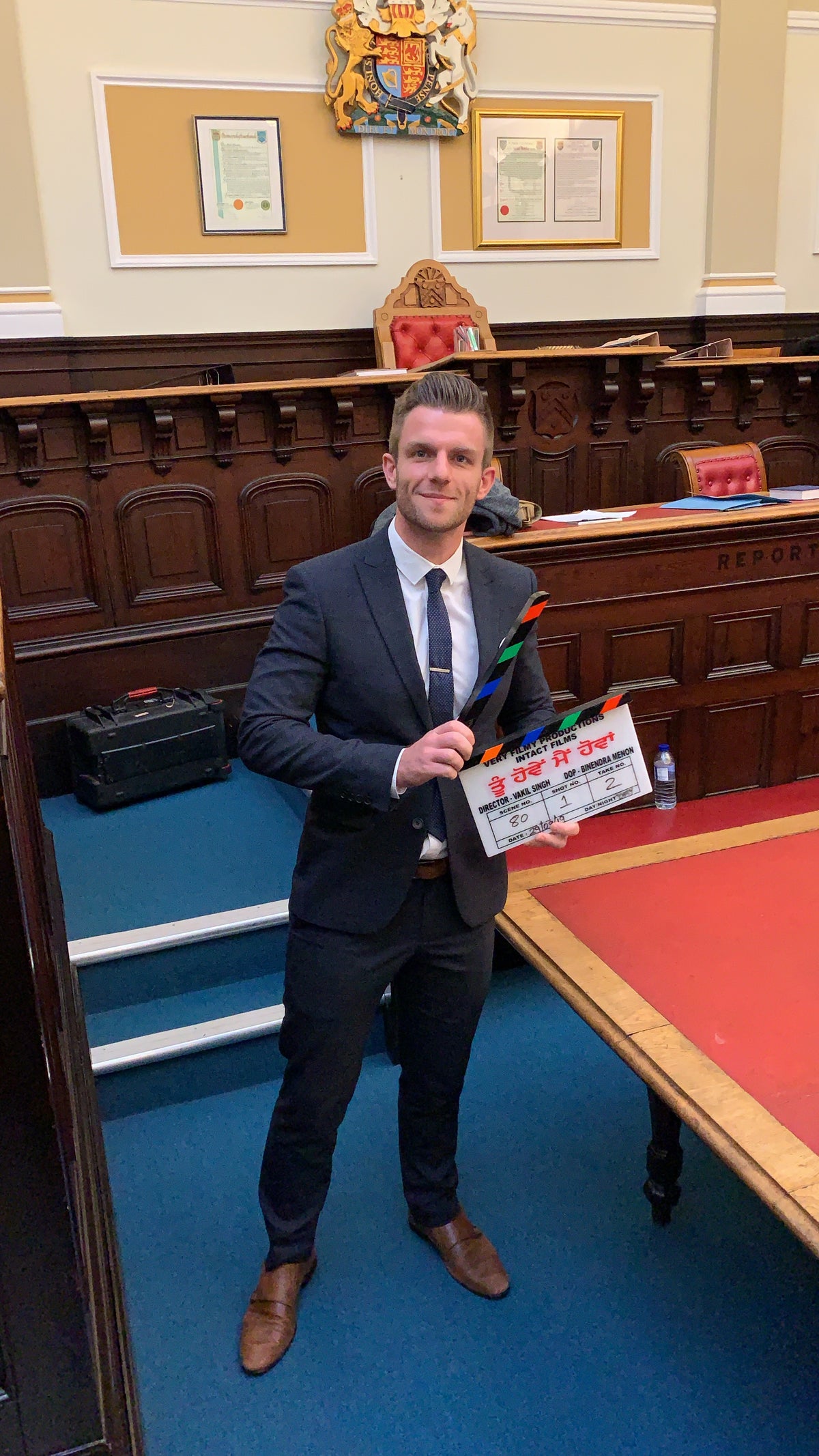 A man in a dark navy suit, white shirt, and tie stands in a courtroom holding a clapperboard with 'The Big Reveal' written on it. The room features dark wood paneling, a blue carpet, and a red table.